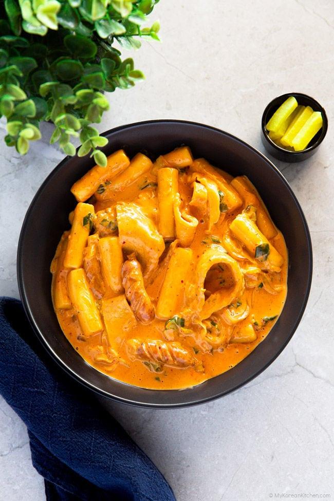 Rose tteokbokki is served in a black pasta bowl, accompanied by slices of yellow pickled radish.